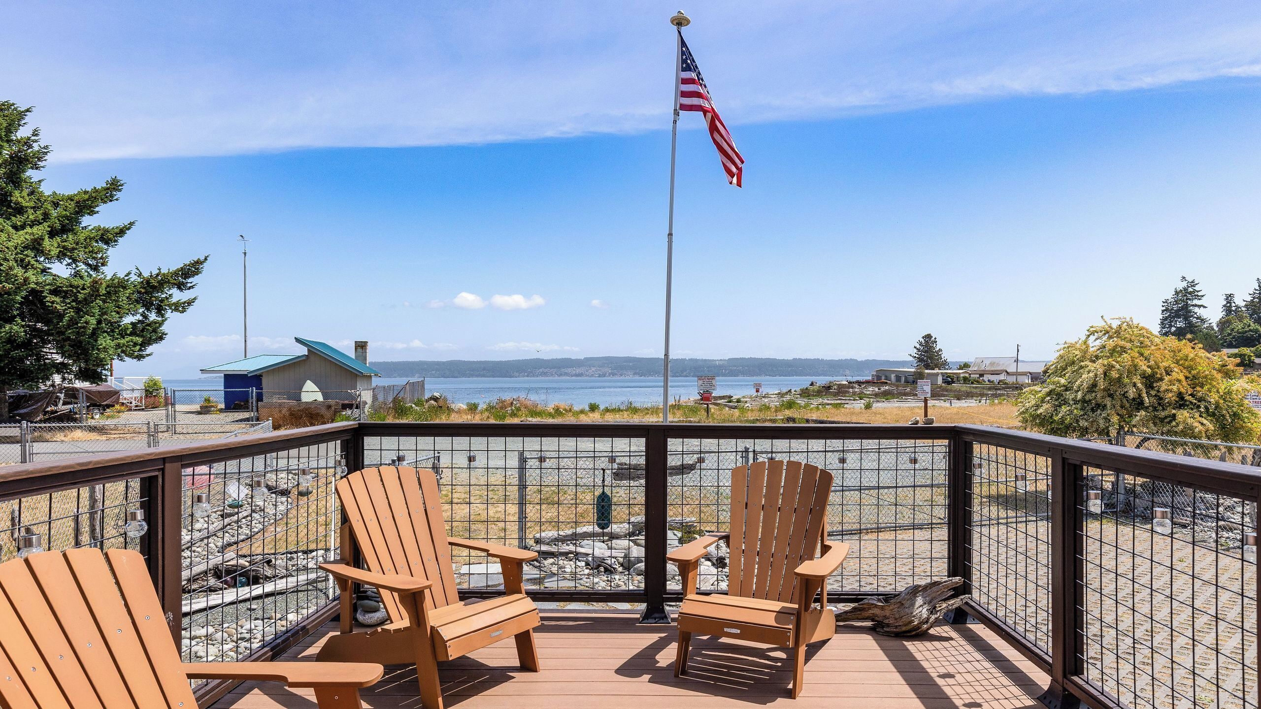 A deck with wooden chairs and a flagpole with an American flag, overlooking a body of water and distant buildings.