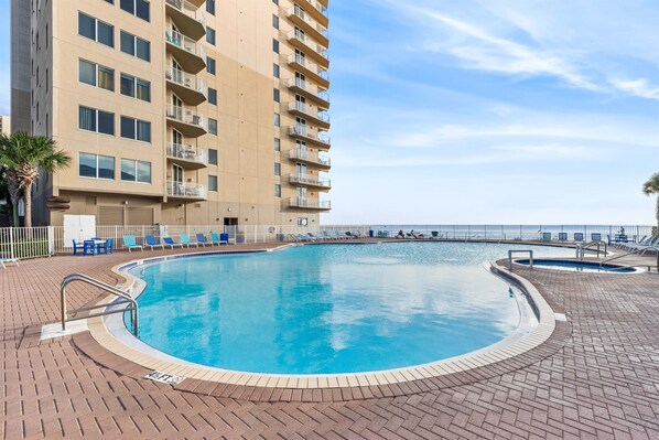 Une piscine entourée d’une terrasse en briques, de chaises longues et d’un grand immeuble doté de balcons.