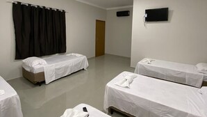 Habitación de hotel con cuatro camas individuales, un televisor de pantalla plana montado en la pared, una lámpara de techo y una puerta que da a otra habitación.
