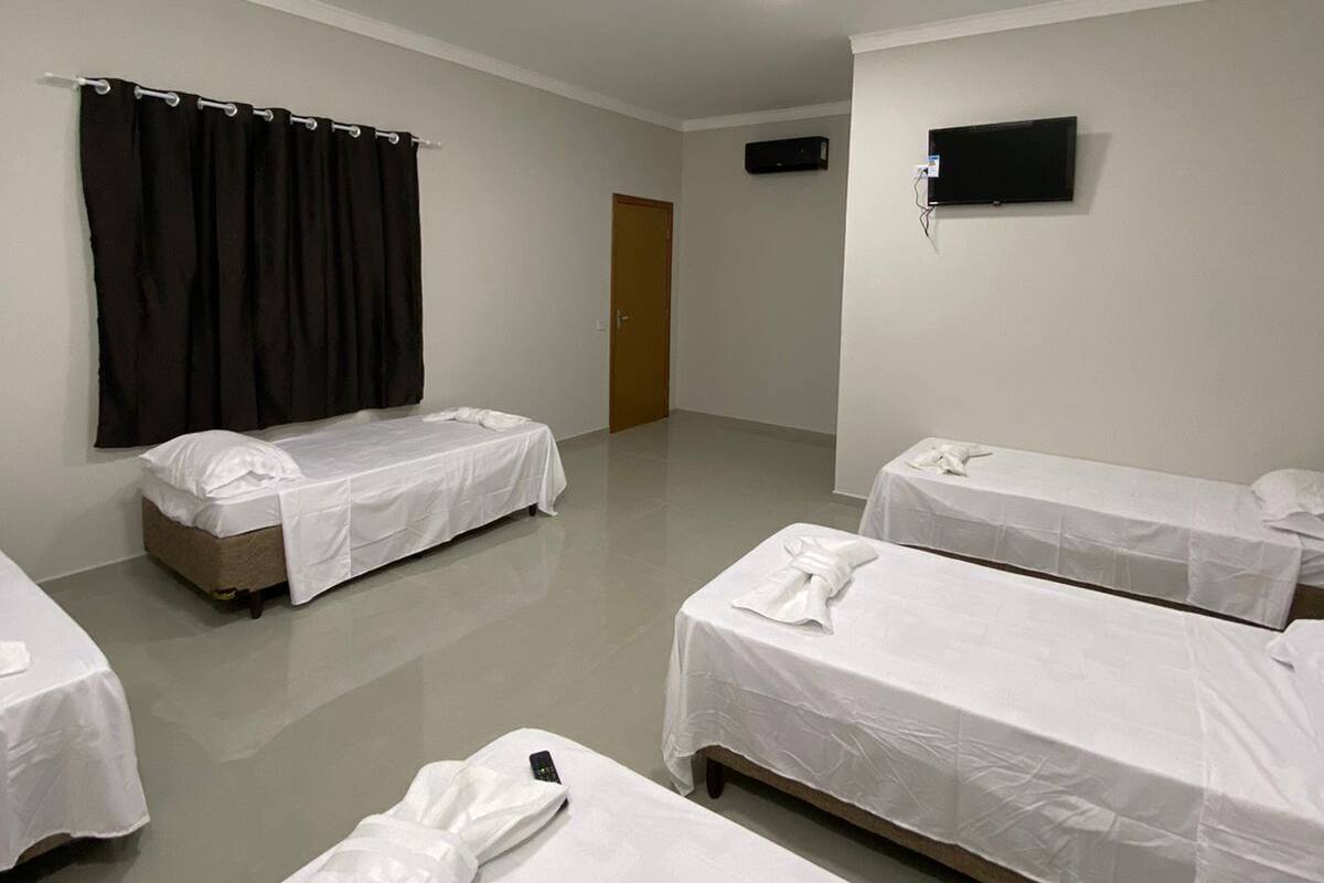 A hotel room with four single beds, a flat-screen TV mounted on the wall, a ceiling light, and a door leading to another room.