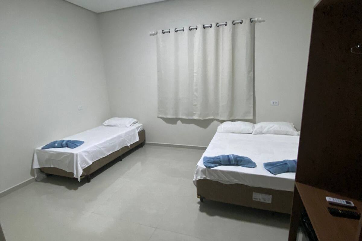 A small, clean room with two single beds, a window with curtains, and a wooden desk with a computer.