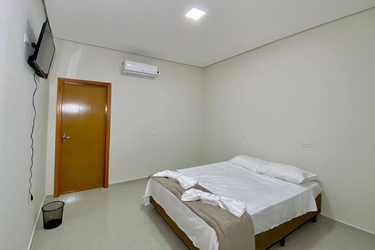 A small, clean room with a single bed, a door, a wall-mounted air conditioner, and a TV on the wall.