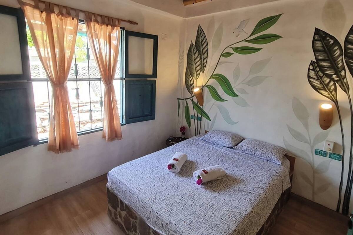 Une chambre à coucher avec un lit, une fenêtre avec des rideaux, une fresque murale et une table de chevet.