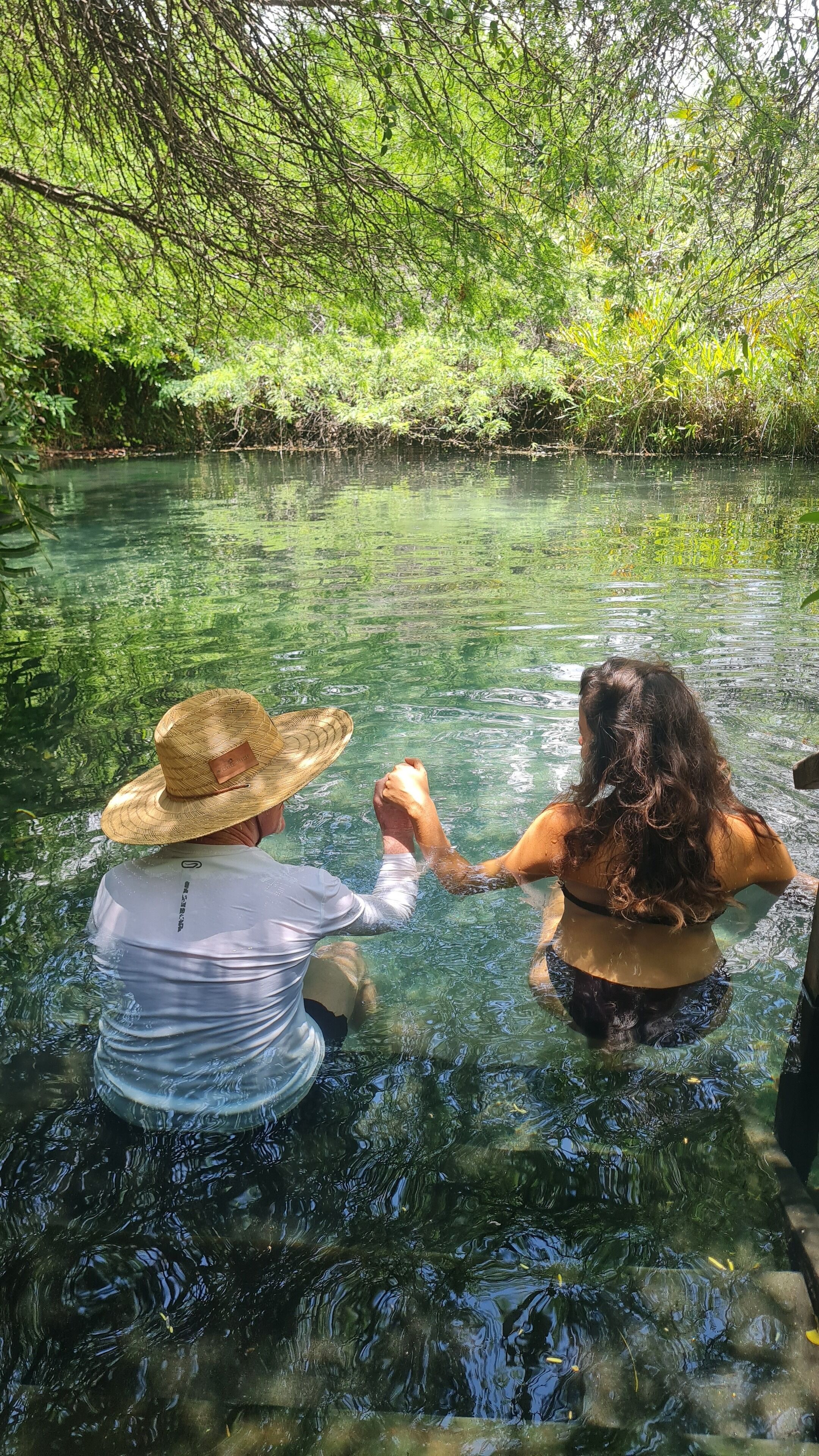 Two people in a clear water body surrounded by dense greenery.