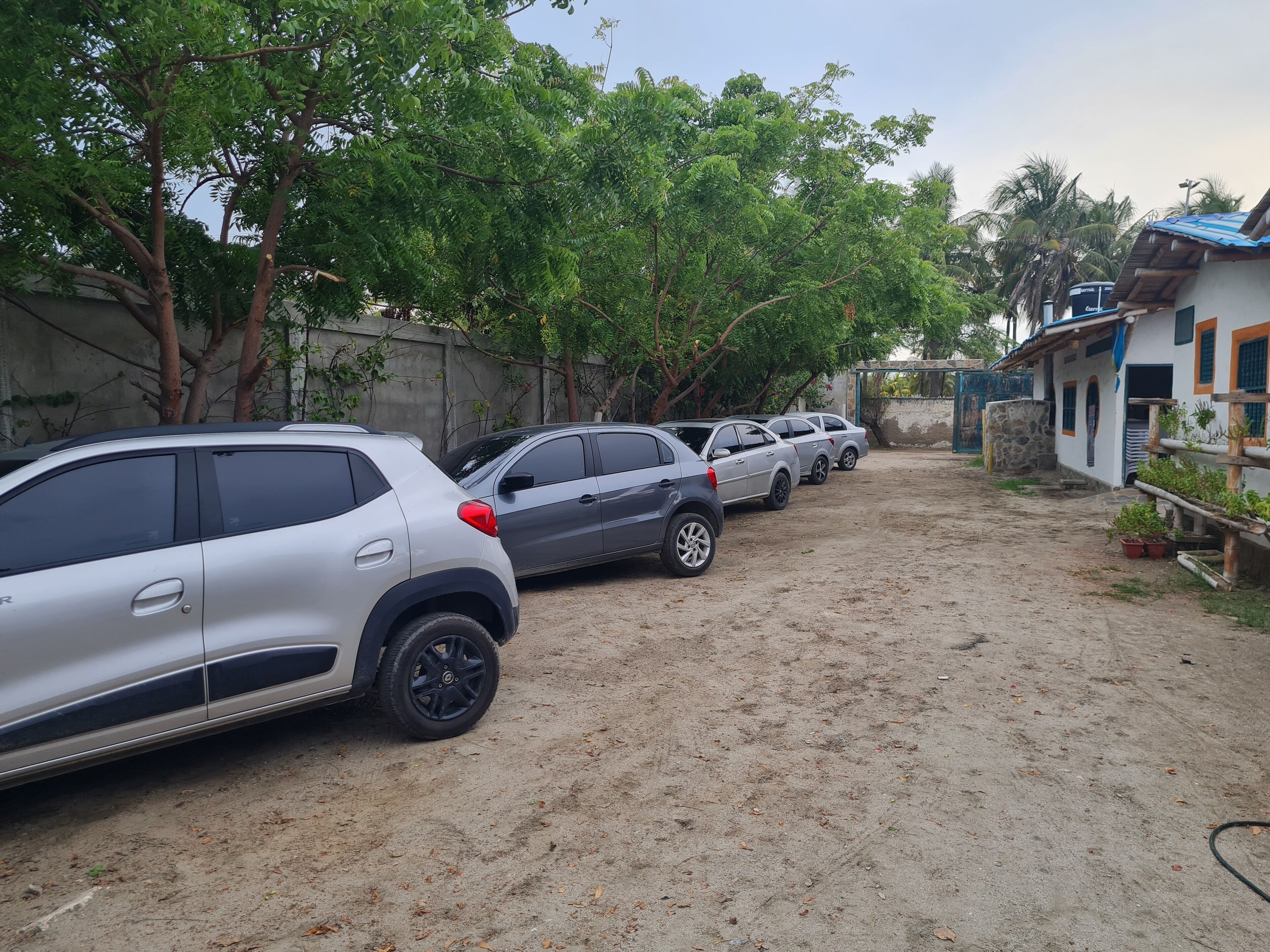 Una fila de coches aparcados en una zona arenosa con árboles y un edificio al fondo.