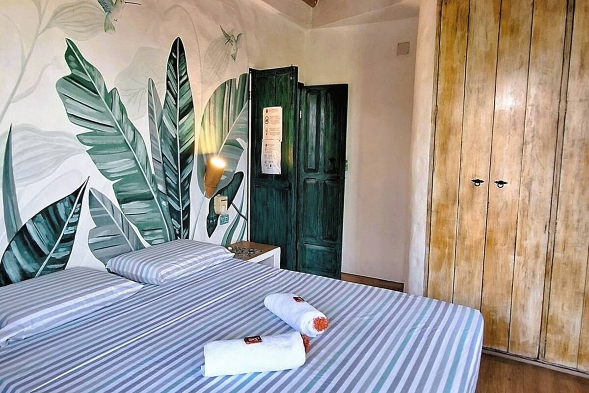 Une chambre avec un lit, une armoire en bois et un mur orné d’une fresque aux motifs tropicaux.
