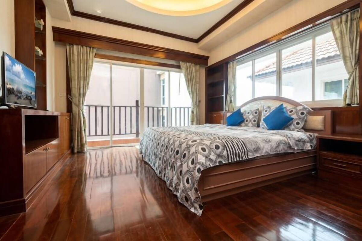 A bedroom with a large bed, wooden flooring, a balcony with curtains, and a TV on a wooden cabinet.