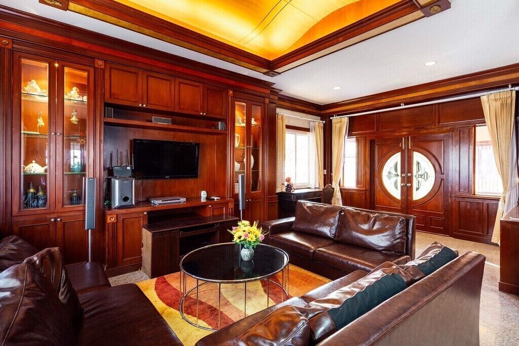 Un salón amplio con un sofá de cuero grande, un televisor de pantalla plana, armarios de madera y un ventanal redondo decorativo.