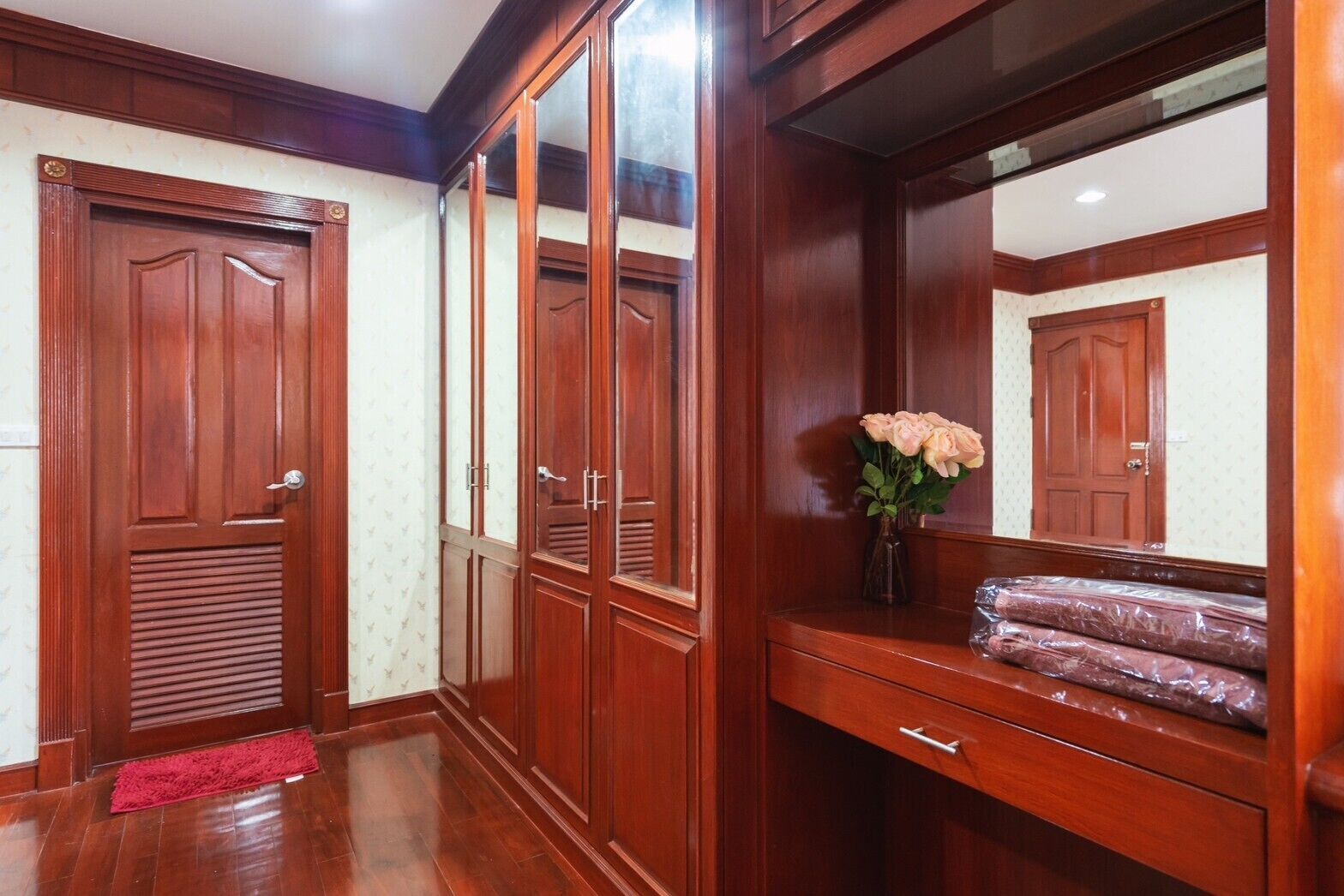 A wooden wardrobe with a mirror, a red mat at the entrance, and a vase with flowers on the vanity.