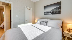 A neatly made bed with white bedding, a nightstand with a lamp, a door, and a wall-mounted painting of a forest scene.
