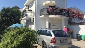 Un edificio de dos plantas con un balcón, un coche aparcado y una persona de pie cerca de la entrada.