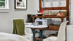 Een gezellige kamer met een groene stoel, een klein rond tafeltje, een witte stoel, een magnetron en een plank met potten en een mand.