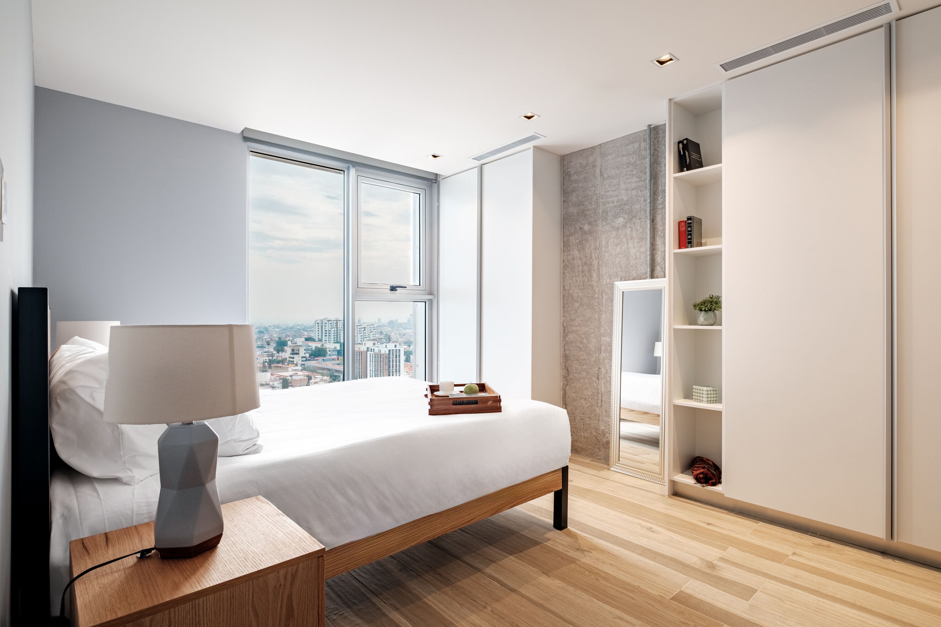 A modern bedroom with a large bed, a bedside table with a lamp, a wardrobe, and a city view through a large window.