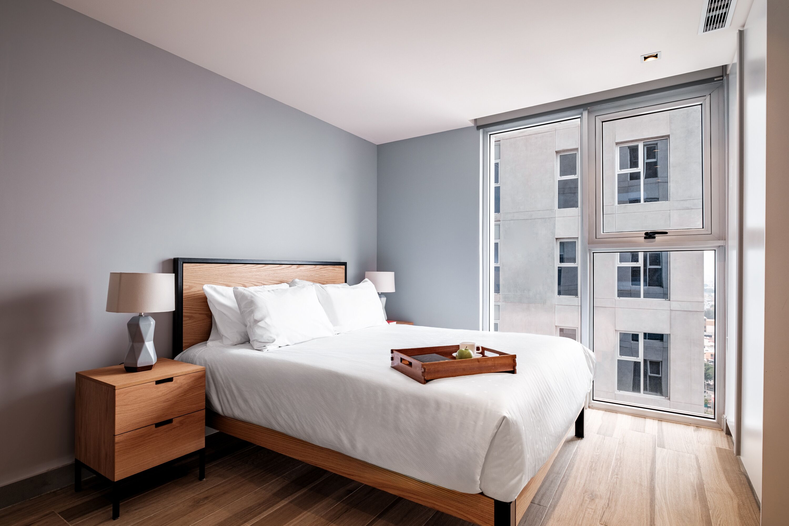 A modern bedroom with a large bed, wooden nightstands, a bedside table with a lamp, and a window with a view of buildings.