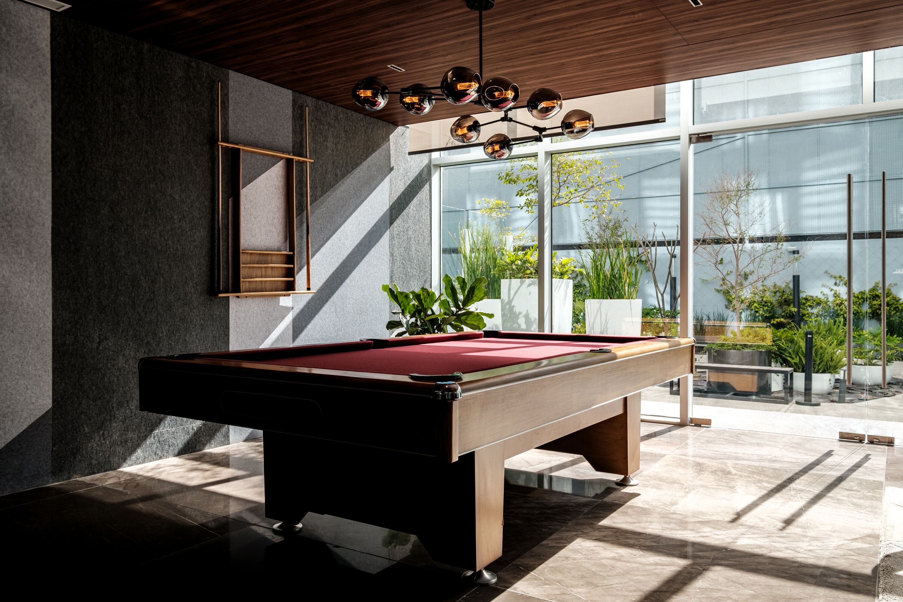 Una mesa de billar en una habitación moderna con ventanas amplias, techo de madera y diseño geométrico en la pared.