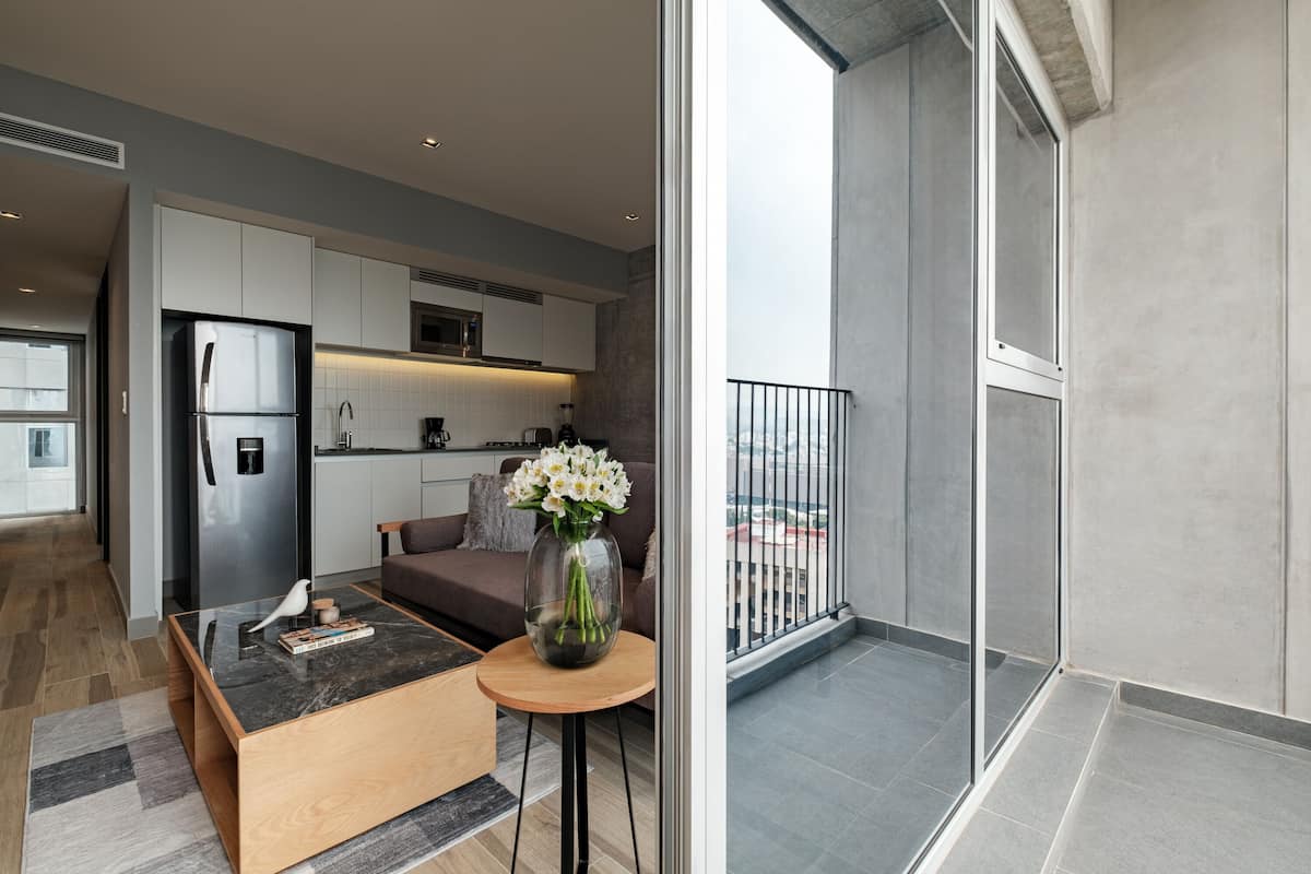 A modern kitchen with a refrigerator, microwave, and coffee maker. There is a dining area with a table and chairs, and a balcony with a railing.