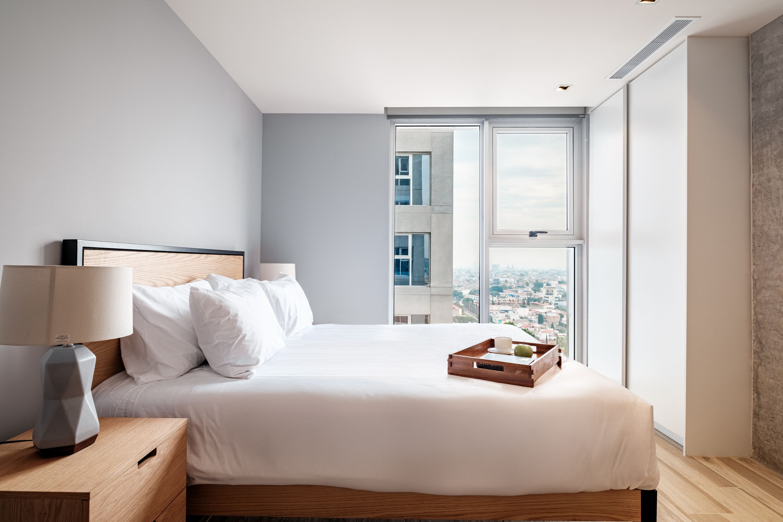 Une chambre d’hôtel moderne avec un grand lit, une table de chevet avec une lampe et une vue sur la ville grâce à de grandes fenêtres.