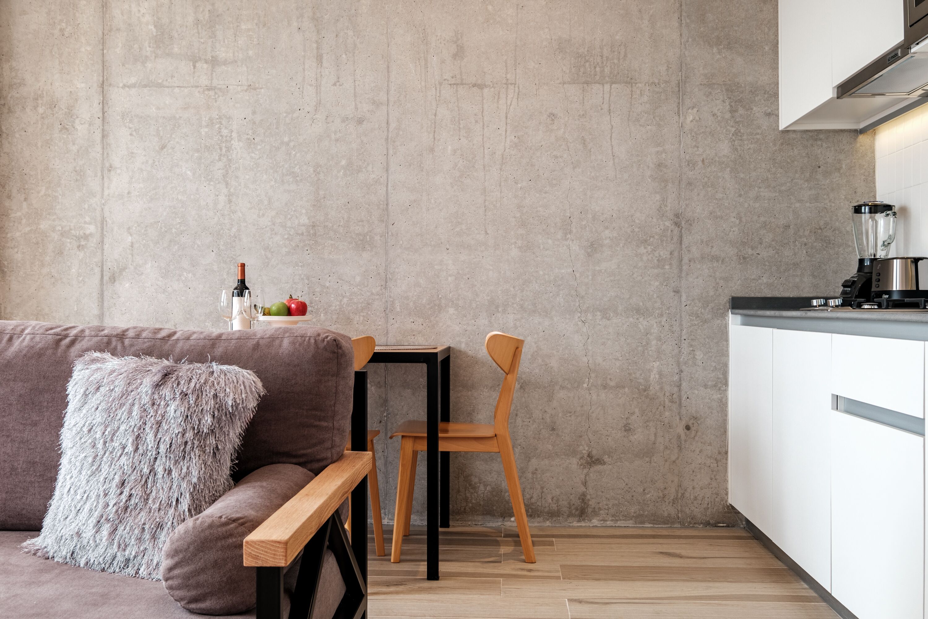 A modern kitchen with a concrete wall, a wooden dining table, and a sofa with a fluffy pillow.