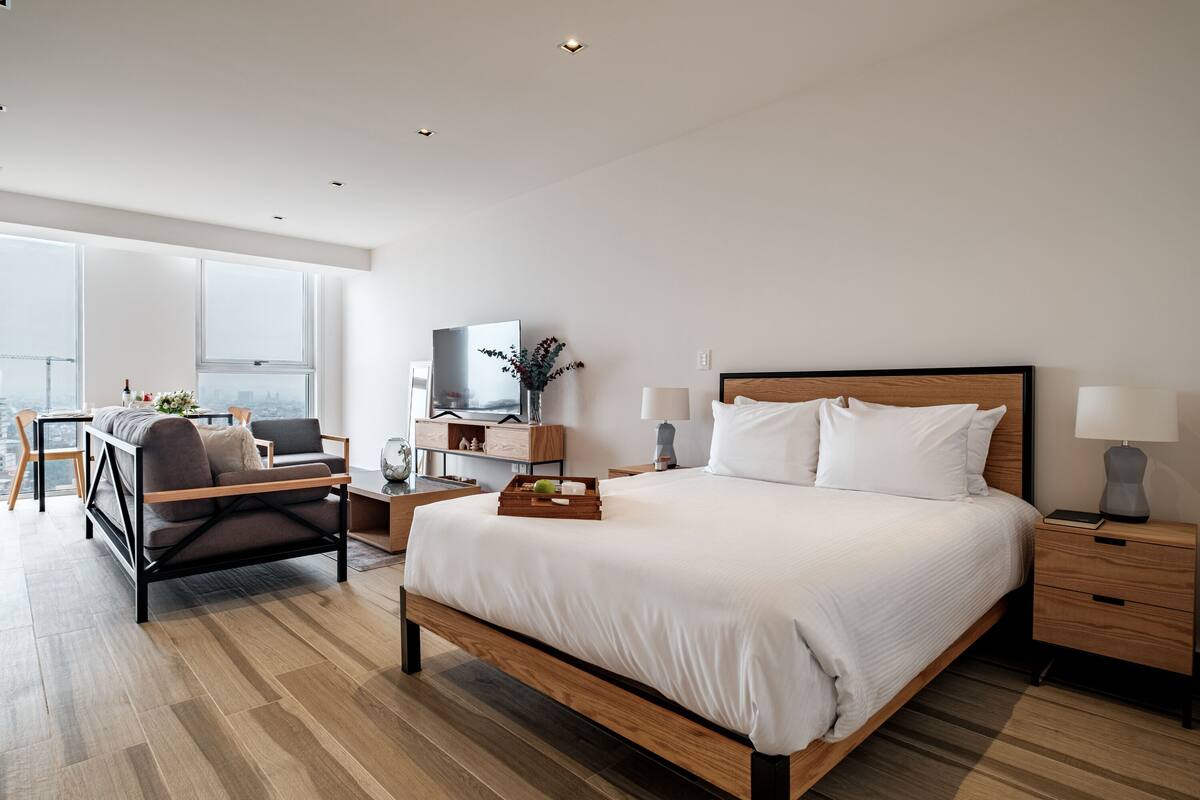 A modern hotel room with a large bed, a wooden nightstand, a bedside table, a desk with a chair, and a large window offering a city view.