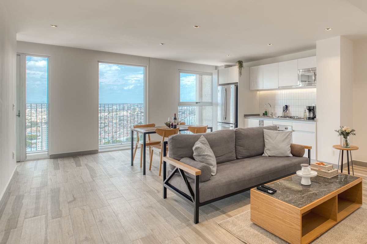 A modern apartment with a living area, dining space, kitchen, and large windows offering city views.
