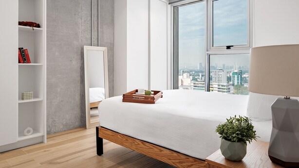 A modern bedroom with a large bed, a bedside table with a lamp, a window with a city view, a wardrobe, and a shelf with books and a tissue box. - Nido GDL (Guadalajara)