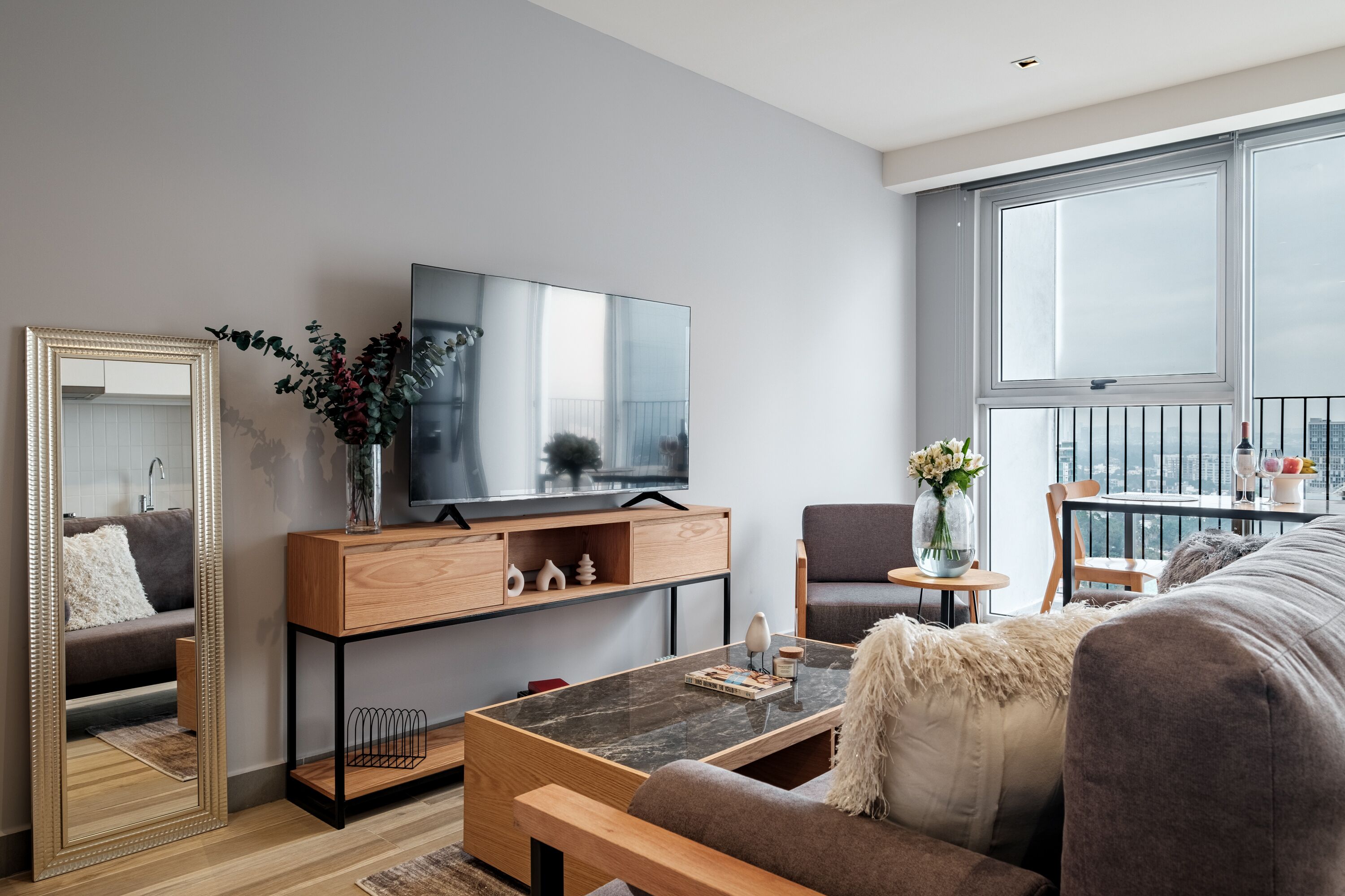 Un salon moderne doté d’un grand téléviseur à écran plat, d’une table basse en bois, d’un canapé gris et d’un balcon offrant une vue sur la ville.