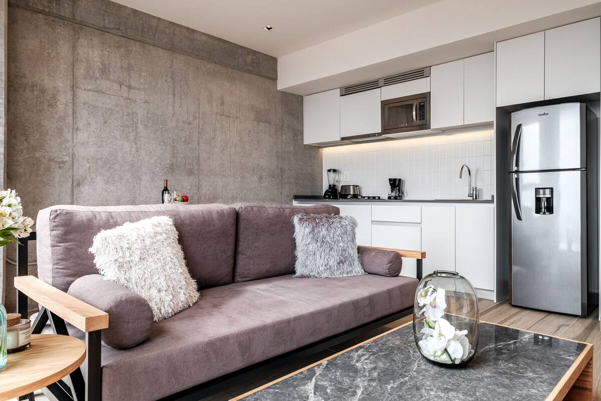 A modern kitchen with a grey sofa, a glass vase with flowers, and a stainless steel refrigerator.