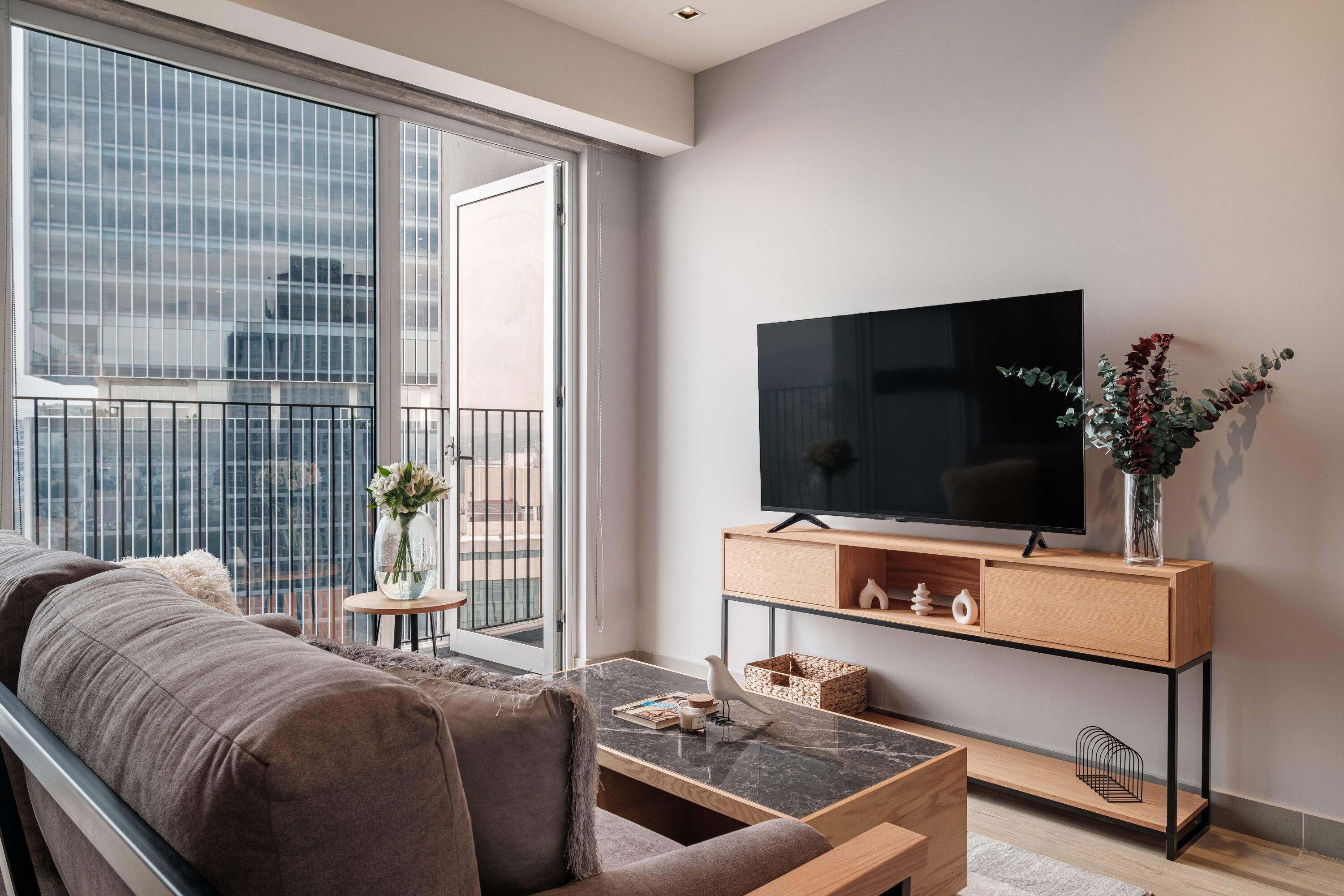 A modern living room with a large window, a flat-screen TV, a sofa, a coffee table, and a small side table with a vase.