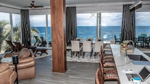 Une chambre d’hôtel moderne dotée d’une grande fenêtre donnant sur la mer, d’un coin repas avec une longue table et d’un espace détente avec des canapés et des fauteuils.