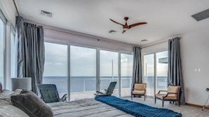 Une chambre d’hôtel avec un grand lit, un ventilateur de plafond et une vue sur la mer grâce à de grandes fenêtres.