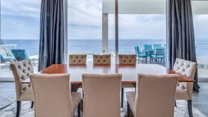 Un espace repas comprenant une table et des chaises, de grandes fenêtres avec des rideaux et une vue sur l’océan.