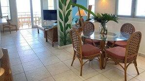 Een eethoek met een glazen tafel, rieten stoelen, een grote plant en een televisie op de achtergrond.