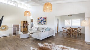 A spacious living area with a sofa, coffee table, and a dining table with chairs. There are two hanging light fixtures and a large abstract painting on the wall.