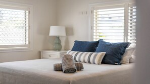 A neatly made bed with a rolled towel, a striped pillow, and a solid blue pillow. There is a bedside table with a lamp and a wall socket.