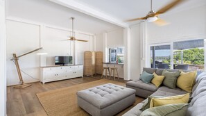A spacious living area with a sofa, ottoman, and a television on a white cabinet.
