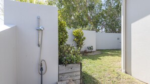Un muro bianco con un tubo di metallo fissato, una fioriera in legno con piante e un prato con alberi sullo sfondo.