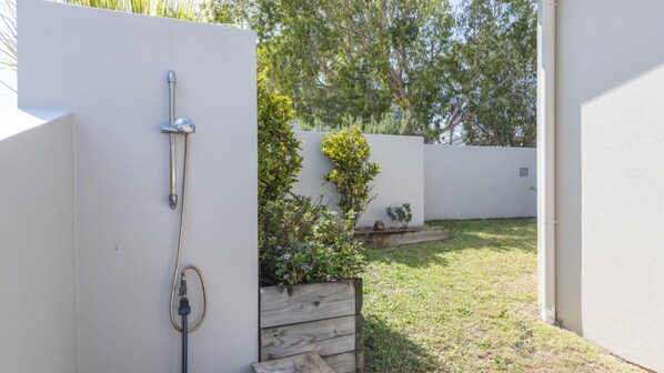A white wall with a metal hose attached to it, a wooden planter box with plants, and a grassy yard with trees in the background.