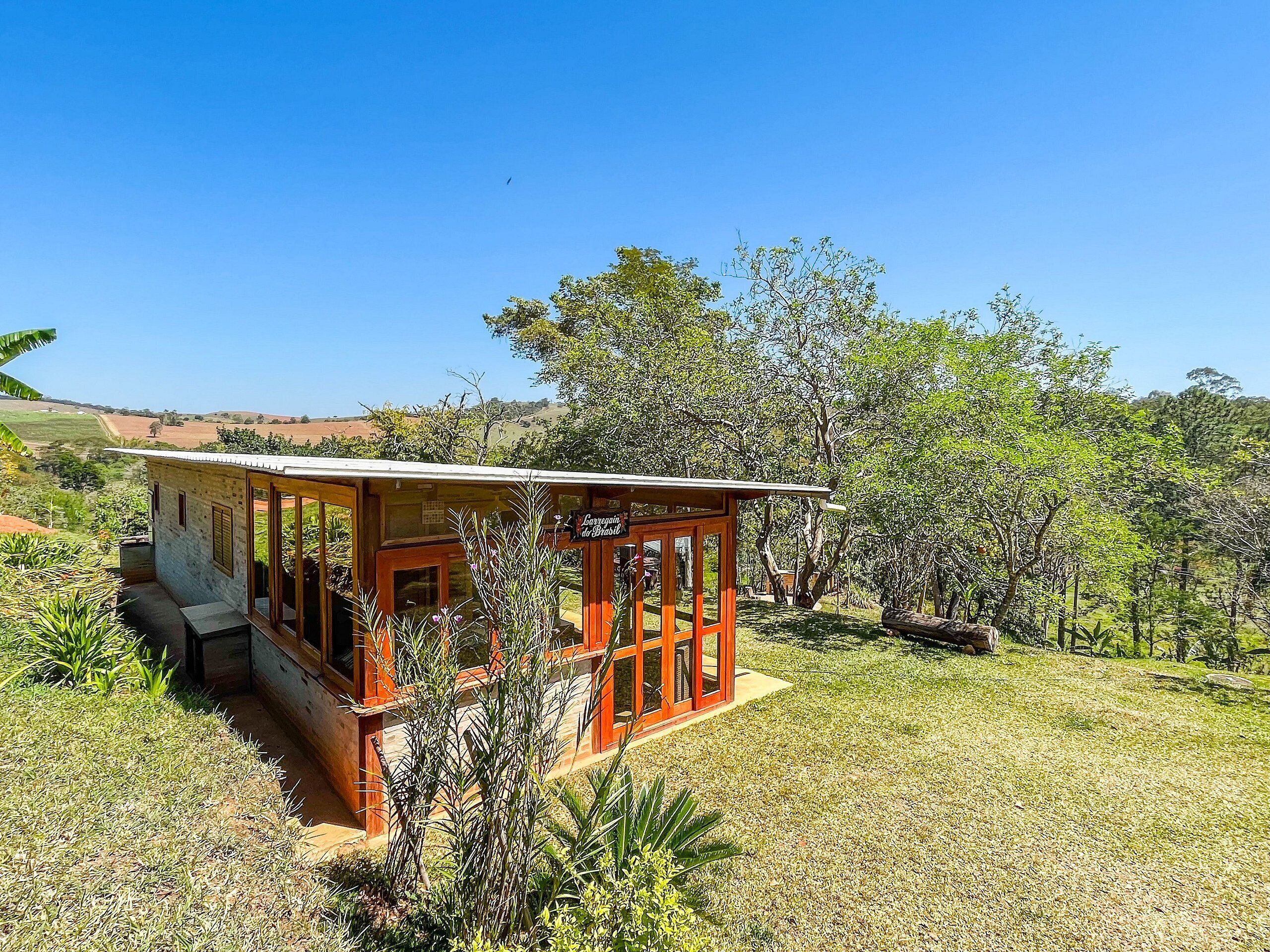 A small wooden structure with a porch, surrounded by grass and trees, with a clear sky in the background.