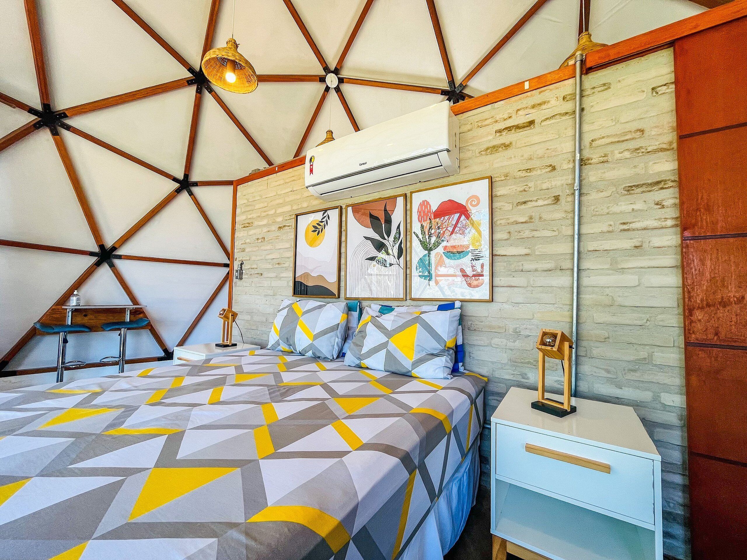 A bedroom with a geometric-patterned bedspread, a white bedside table, a wall-mounted air conditioner, and a wooden bench.