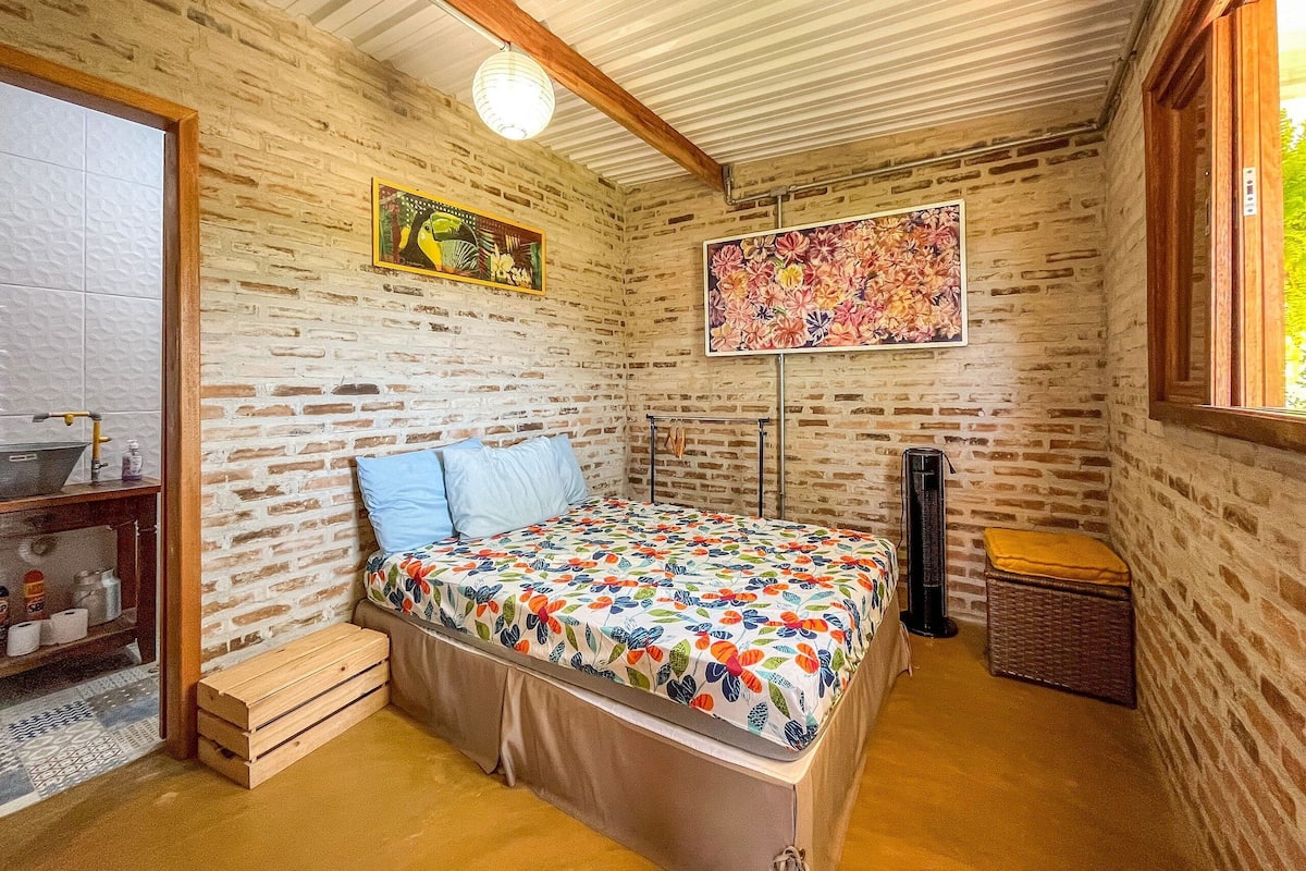 A bedroom with a bed, a wooden bench, a small table with toiletries, a wall-mounted heater, and a window with a view of greenery.