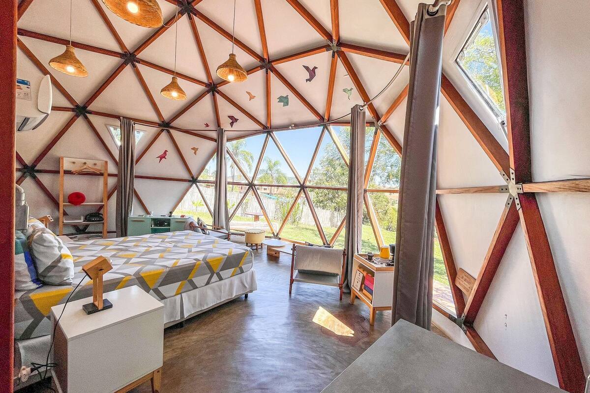 A spacious room with a large bed, a desk, and a chair, featuring a geometric ceiling design and large windows.