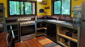 A kitchen with a gas stove, oven, sink, and a water dispenser. There are two windows with decorative grills, a colorful backsplash, and a wooden ceiling. - Amazing Jungle Retreat Close to San Ignacio (Bullet Tree Falls)