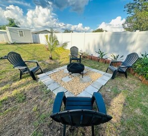 Een achtertuin met een vuurkorf, vier Adirondack stoelen en een kleine palmboom.