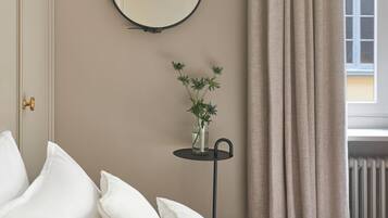 A bedroom with a bed, pillows, a round mirror, a small table with a plant, and a window with curtains.