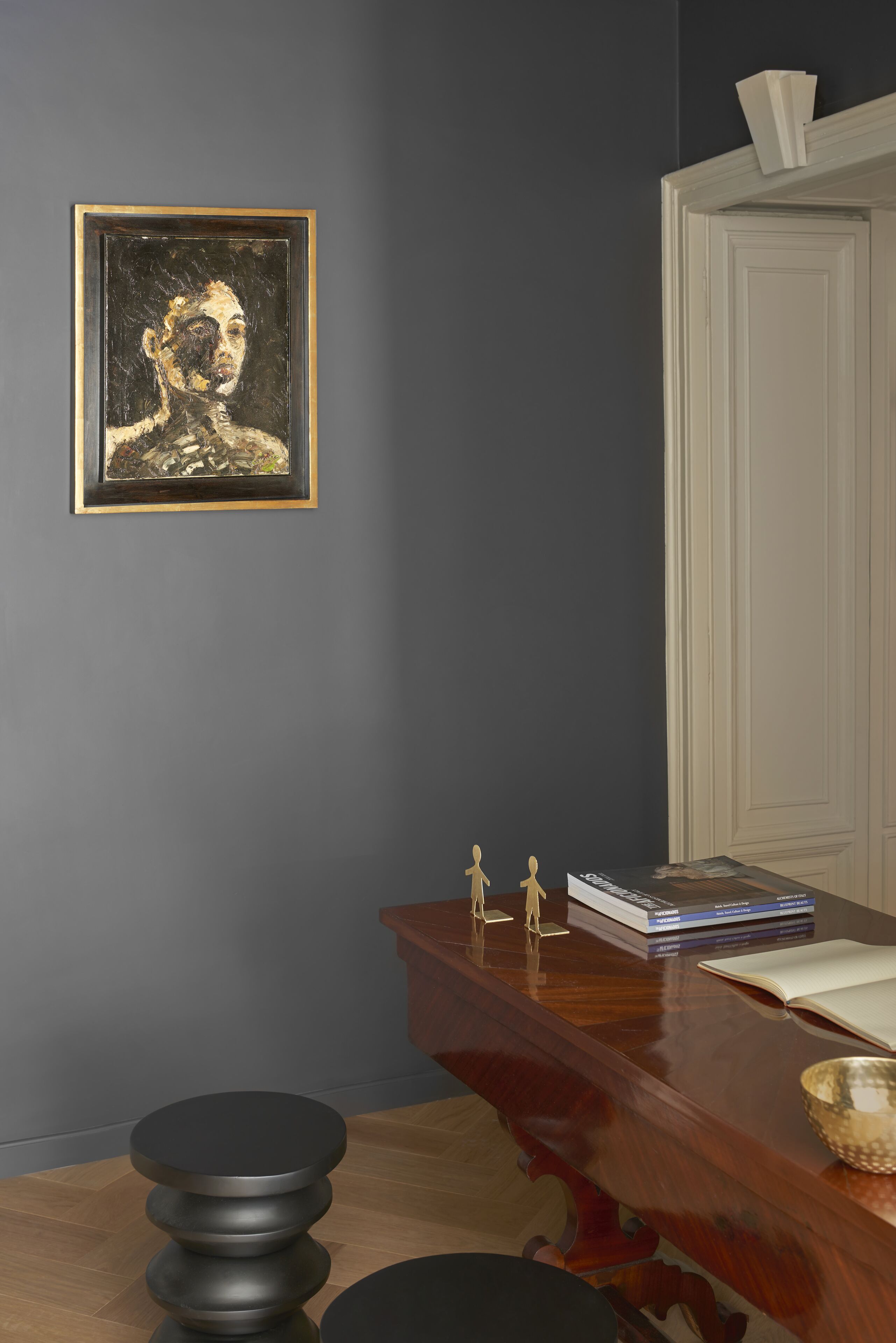 Una habitación con pared gris oscuro, un cuadro enmarcado, un escritorio de madera con libros y un cuenco, y una puerta blanca.