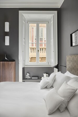 Un dormitorio con una cama grande, una ventana con vistas a edificios, una mesita de noche con una lámpara y un cabecero con un diseño estampado.
