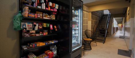 A vending machine stocked with snacks and drinks, located in a hallway with a staircase and a chair.