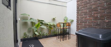 A small patio area with a wooden table and chairs, a black trash bin, and large green plants against a white wall with a brick accent.