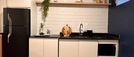 Una cucina compatta con mobili bianchi, un frigorifero nero, uno scaffale in legno con oggetti decorativi e una piccola pianta in vaso.
