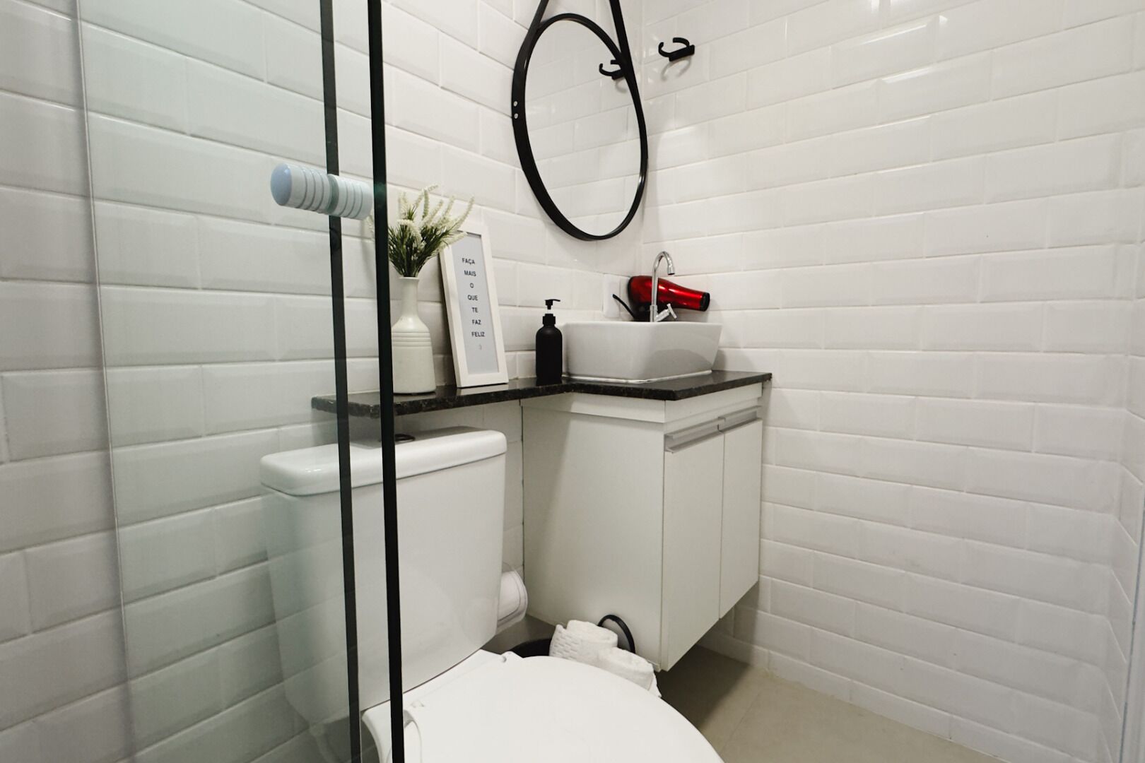 Un baño moderno con azulejos blancos tipo metro, un inodoro blanco, un lavamanos con encimera negra, un secador rojo y un espejo circular negro.